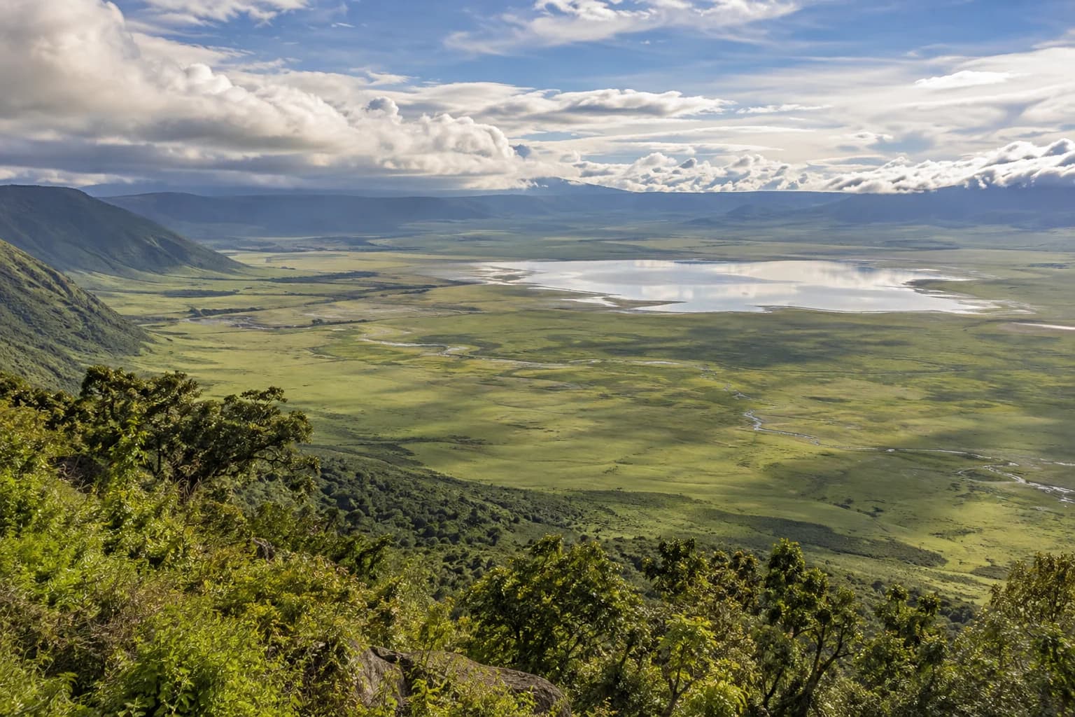 AI-generated illustration: Panoramic view down from Ngorongoro crater rim into the green caldera floor, a distant reflective lake beneath low clouds, natural landscape without close animals.
